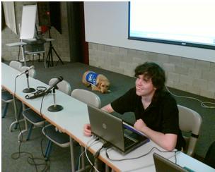 Photo de Cédric dans une salle de conférence assit derrière une table devant son ordinateur. Neptune est sur un podium derrière, et il le regarde. 